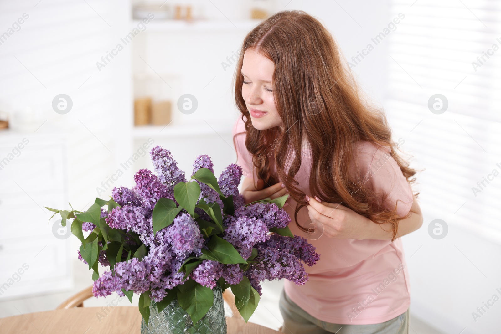 Beautiful teenage girl sniffing lilac flowers at table indoors Photo of Beautiful teenage girl sniffing lilac flowers at table indoors