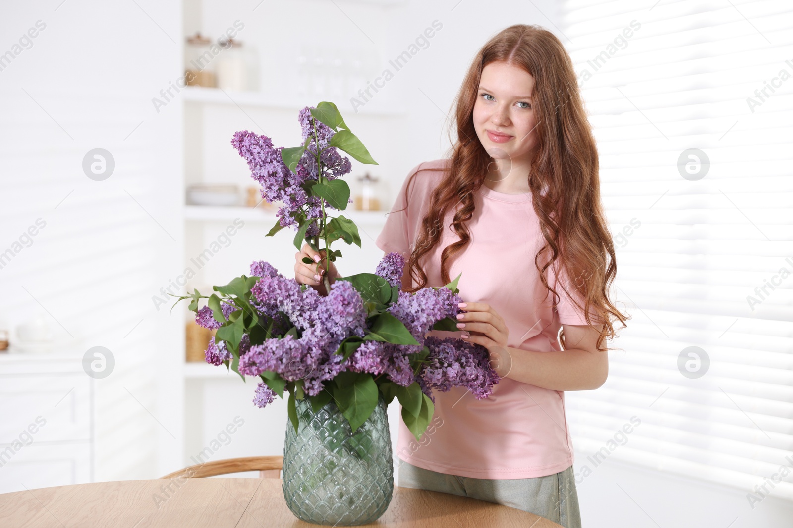 Beautiful teenage girl putting branch with lilac flowers into vase at table indoors Photo of Beautiful teenage girl putting branch with lilac flowers into vase at table indoors