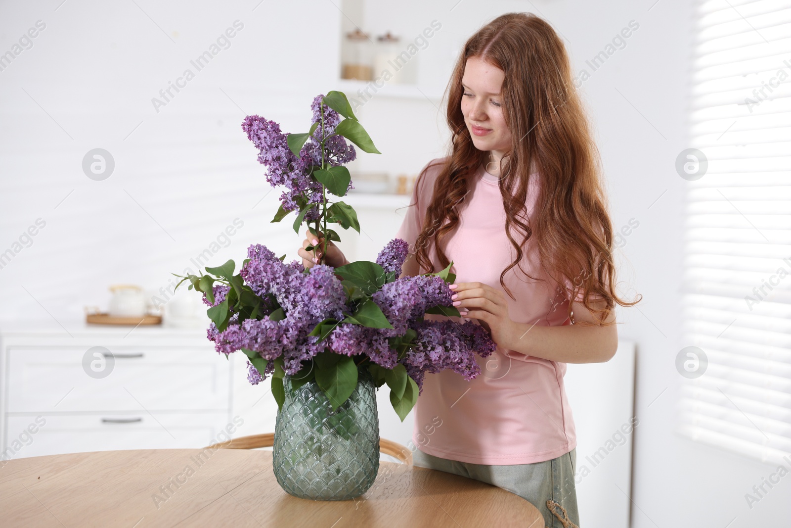 Beautiful teenage girl putting branch with lilac flowers into vase at table indoors Photo of Beautiful teenage girl putting branch with lilac flowers into vase at table indoors
