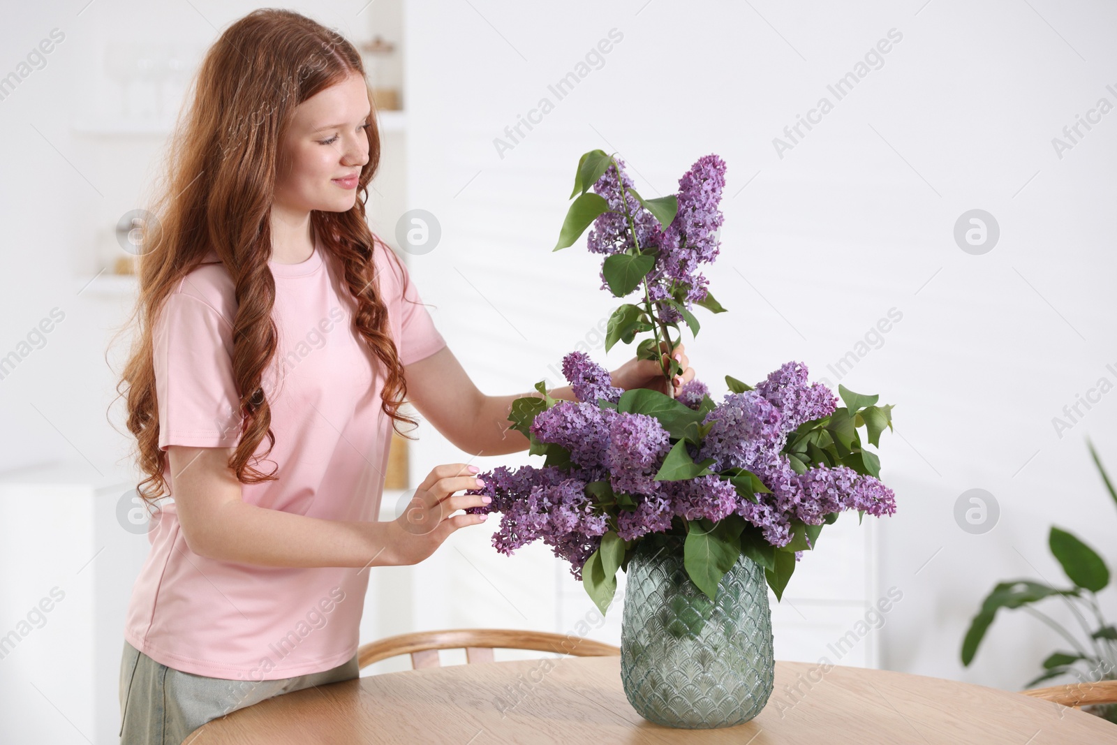 Beautiful teenage girl putting branch with lilac flowers into vase at table indoors Photo of Beautiful teenage girl putting branch with lilac flowers into vase at table indoors