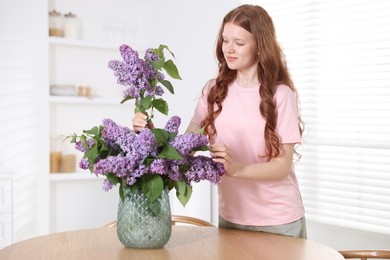 Photo of Beautiful teenage girl putting branch with lilac flowers into vase at table indoors
