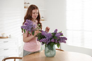 Photo of Beautiful teenage girl putting branch with lilac flowers into vase at table indoors