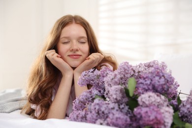 Photo of Beautiful teenage girl with bouquet of lilac flowers on bed indoors