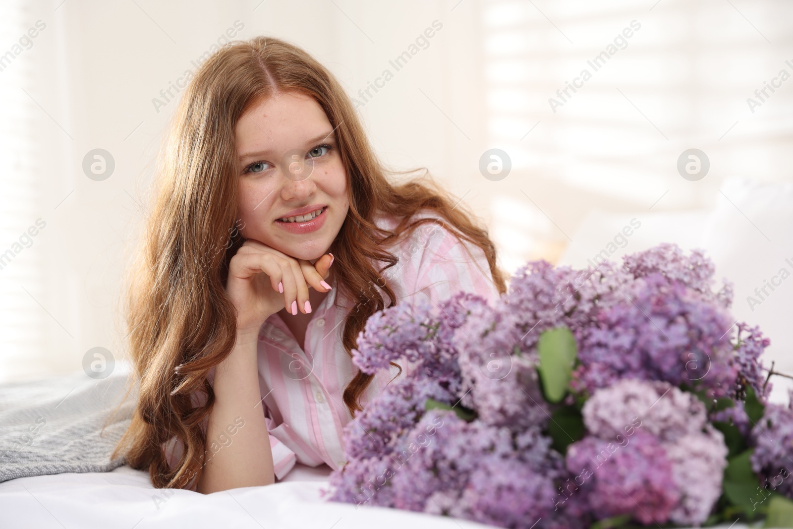 Smiling teenage girl with bouquet of lilac flowers on bed indoors Photo of Smiling teenage girl with bouquet of lilac flowers on bed indoors