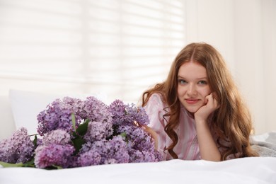Photo of Beautiful teenage girl with bouquet of lilac flowers on bed indoors