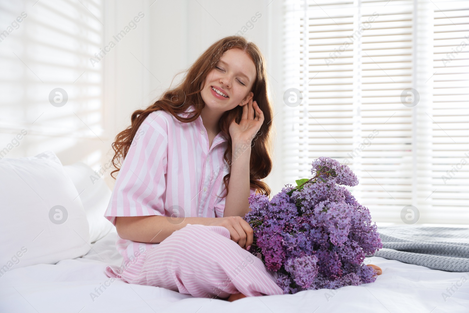Smiling teenage girl with bouquet of lilac flowers on bed indoors Photo of Smiling teenage girl with bouquet of lilac flowers on bed indoors