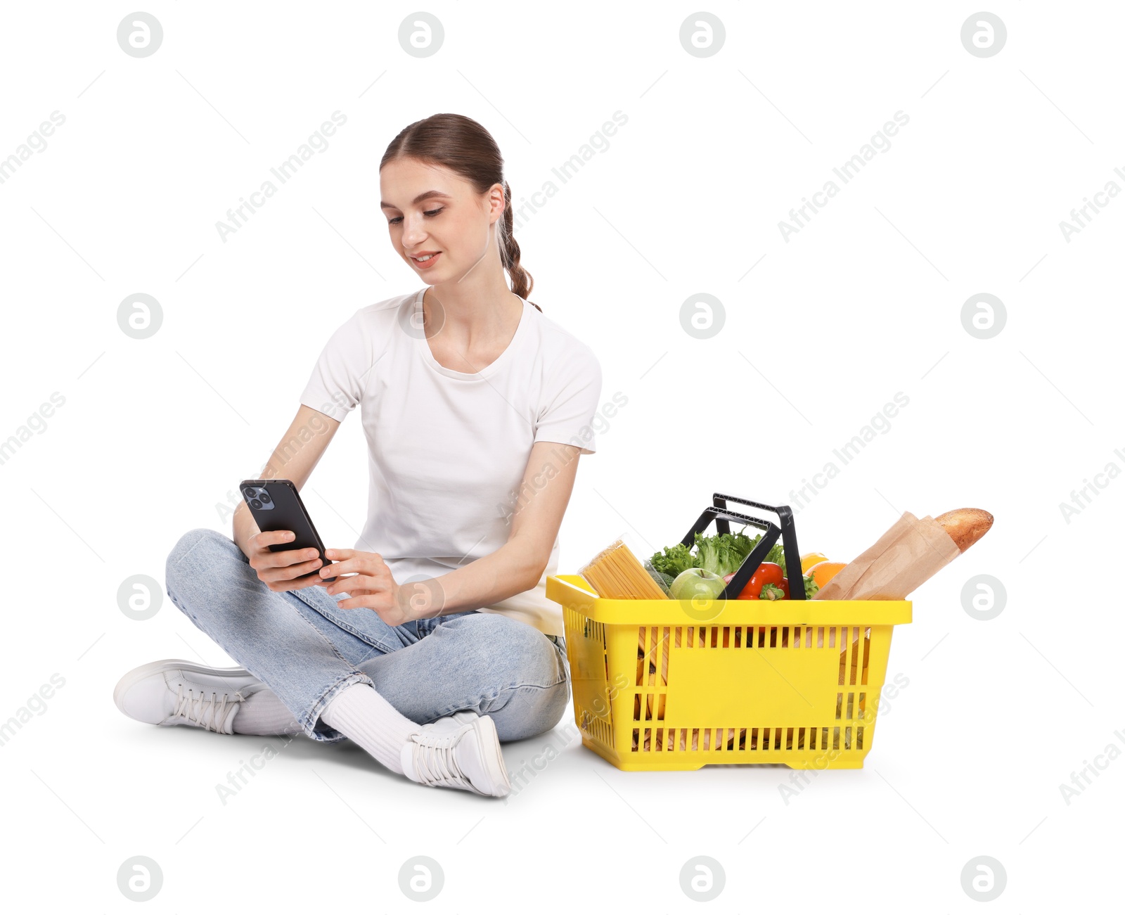 Woman with shopping basket full of products and smartphone on white background Photo of Woman with shopping basket full of products and smartphone on white background