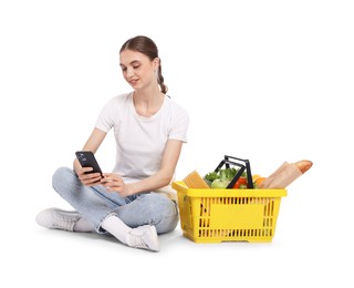 Photo of Woman with shopping basket full of products and smartphone on white background