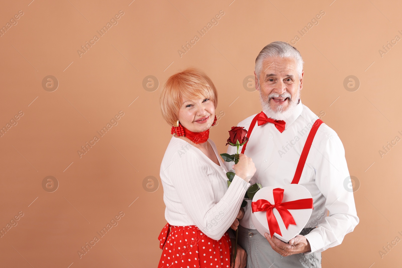 Lovely senior couple with gift box and rose on light brown background, space for text Photo of Lovely senior couple with gift box and rose on light brown background, space for text