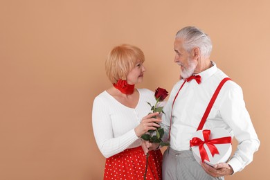 Photo of Lovely senior couple with gift box and rose on light brown background, space for text