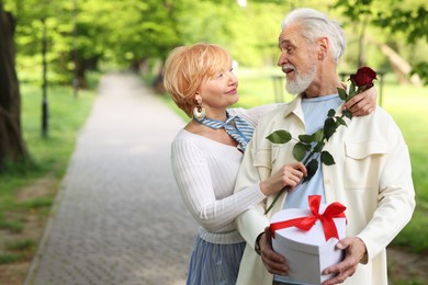 Photo of Lovely senior couple with gift box and rose in park, space for text