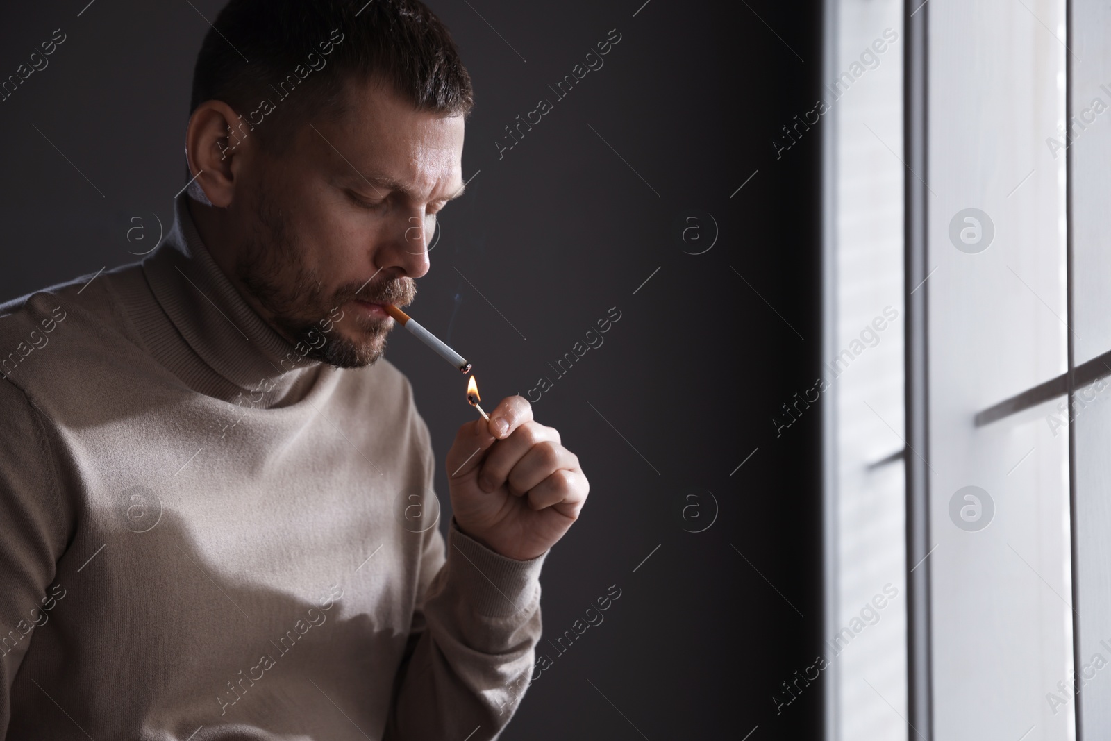 Man lighting cigarette with match near window indoors, space for text Photo of Man lighting cigarette with match near window indoors, space for text