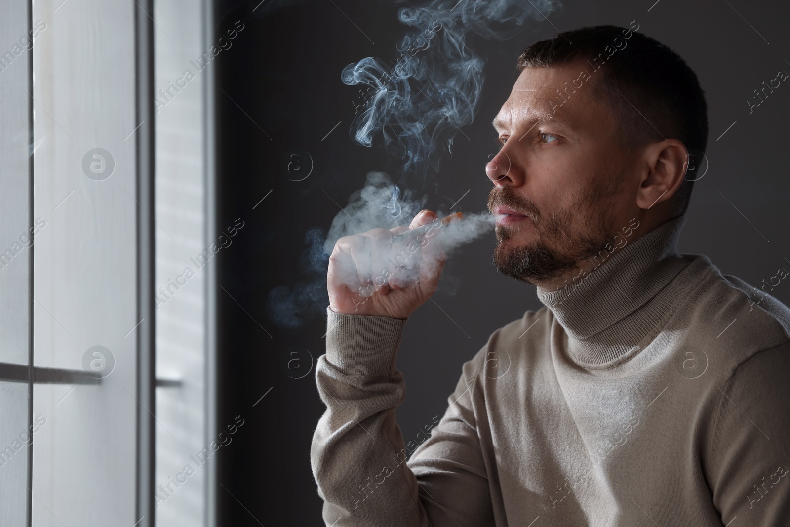 Man smoking cigarette near window indoors, space for text Photo of Man smoking cigarette near window indoors, space for text