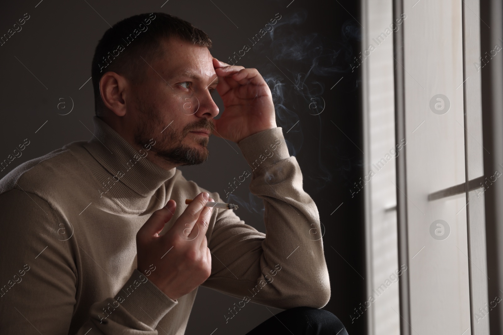 Man smoking cigarette near window in room Photo of Man smoking cigarette near window in room