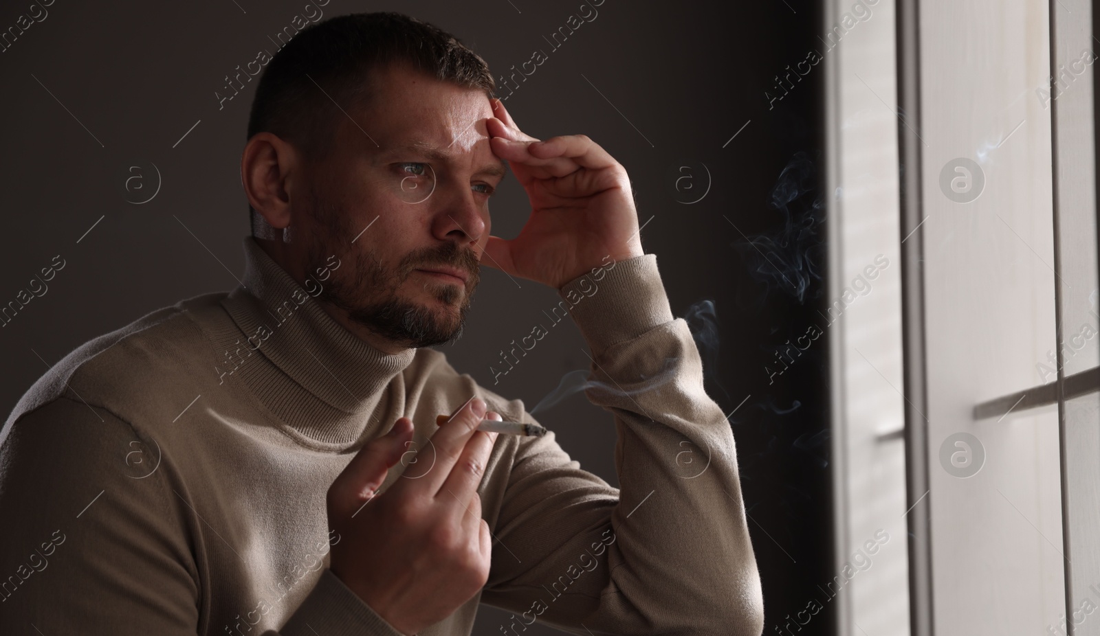 Man smoking cigarette near window in room Photo of Man smoking cigarette near window in room