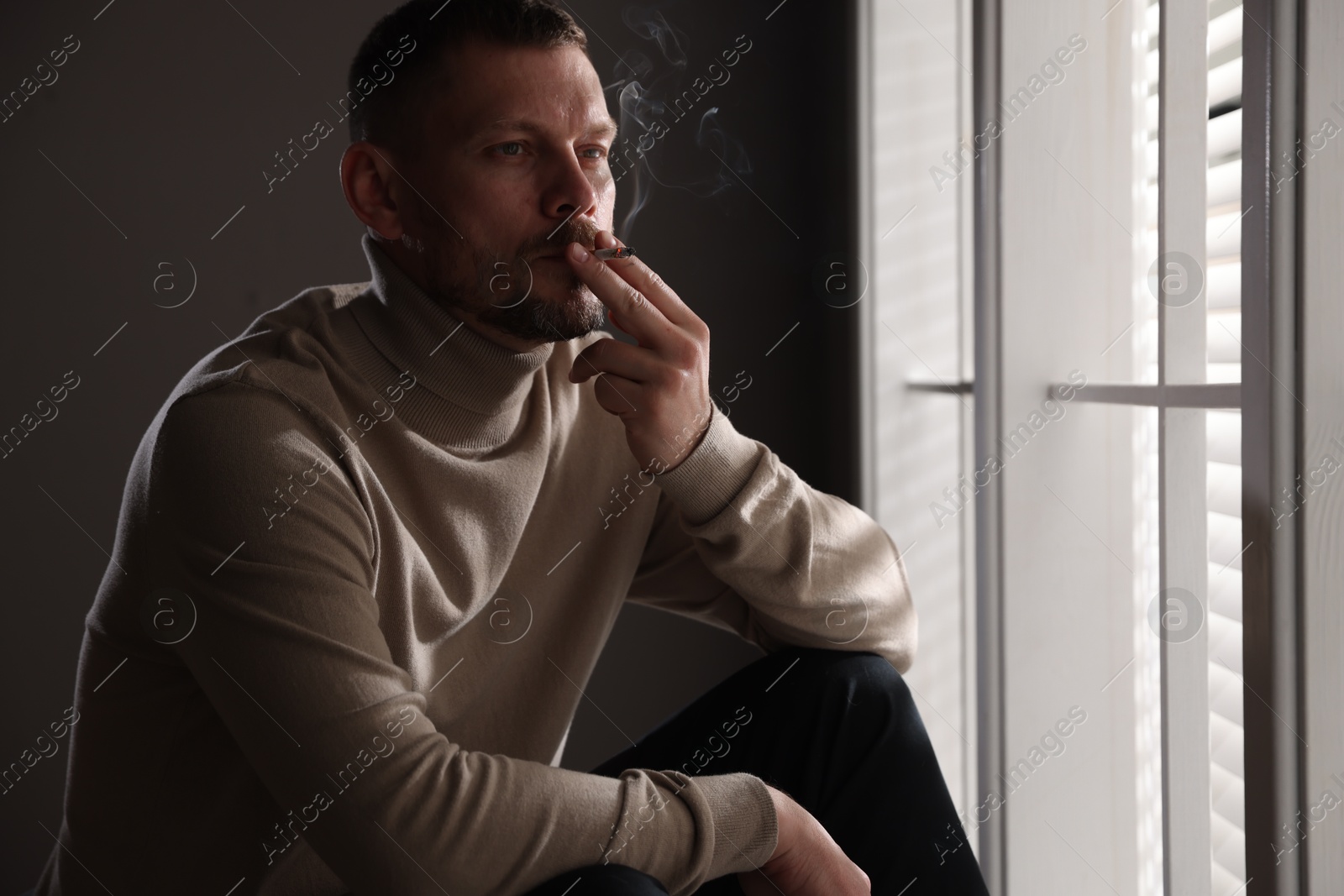 Man smoking cigarette near window in room Photo of Man smoking cigarette near window in room