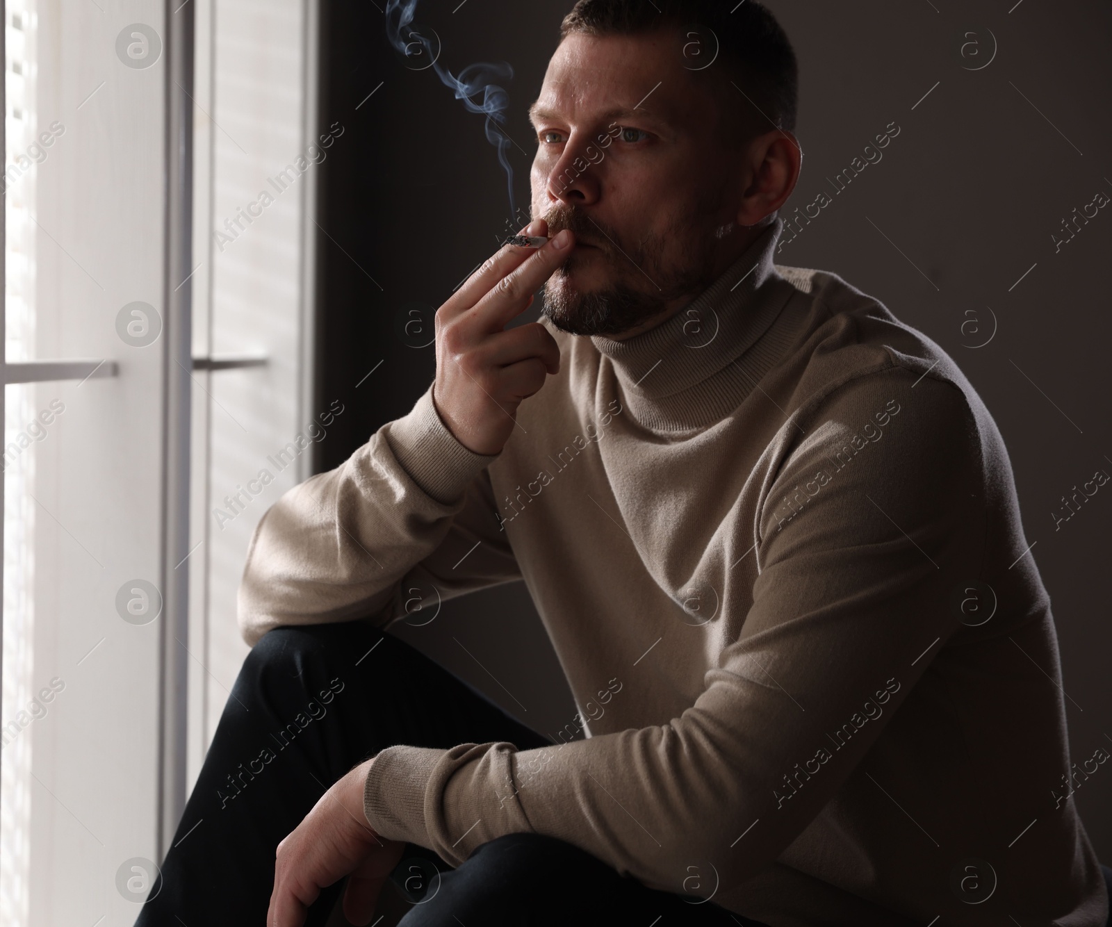 Man smoking cigarette near window in room Photo of Man smoking cigarette near window in room