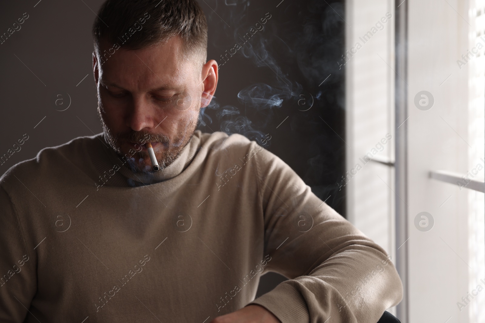 Man smoking cigarette near window in room Photo of Man smoking cigarette near window in room
