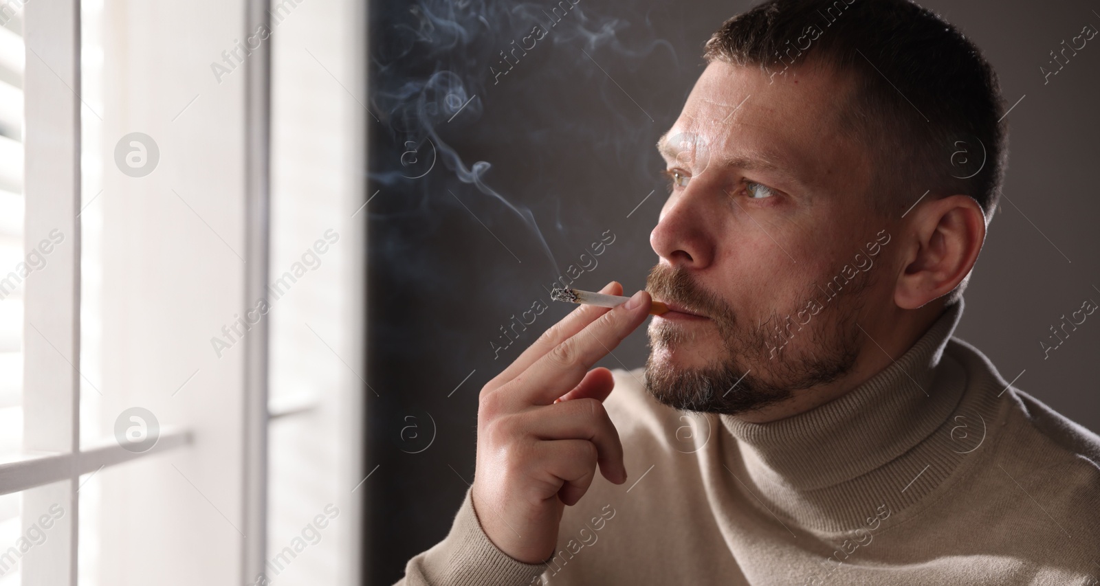 Man smoking cigarette near window indoors, space for text Photo of Man smoking cigarette near window indoors, space for text