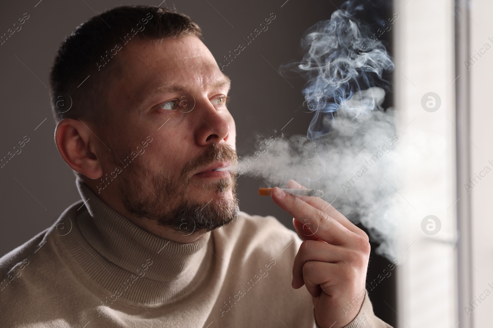 Man smoking cigarette near window in room Photo of Man smoking cigarette near window in room