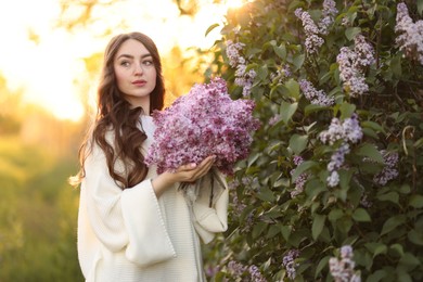 Photo of Beautiful woman with bouquet of lilac flowers near bush outdoors in morning