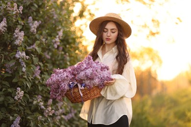 Photo of Beautiful woman with basket of lilac flowers near bush outdoors in morning