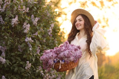 Photo of Smiling woman with basket of lilac flowers near bush outdoors in morning