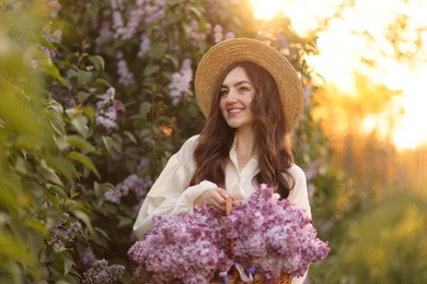 Photo of Smiling woman with basket of lilac flowers near bush outdoors in morning