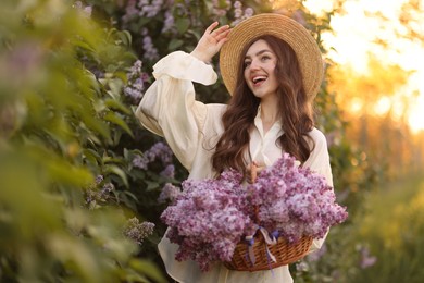 Photo of Happy woman with basket of lilac flowers near bush outdoors in morning