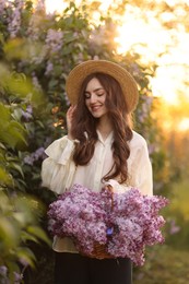 Photo of Smiling woman with basket of lilac flowers near bush outdoors in morning