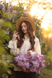 Photo of Beautiful woman with basket of lilac flowers near bush outdoors in morning