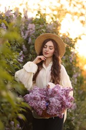 Photo of Beautiful woman with basket of lilac flowers near bush outdoors in morning