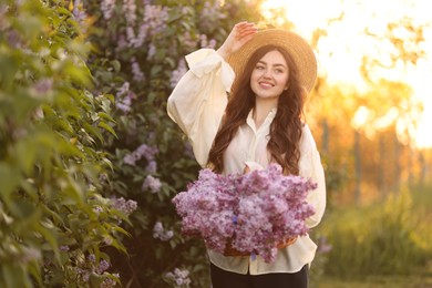 Photo of Smiling woman with basket of lilac flowers near bush outdoors in morning