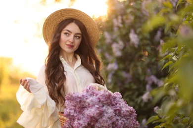Photo of Beautiful woman with basket of lilac flowers near bush outdoors in morning
