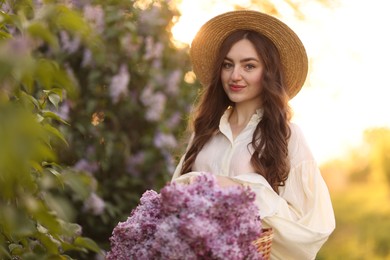 Photo of Beautiful woman with basket of lilac flowers near bush outdoors in morning
