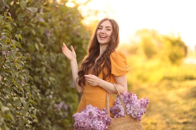 Photo of Smiling woman with bag of lilac flowers near bush outdoors in morning