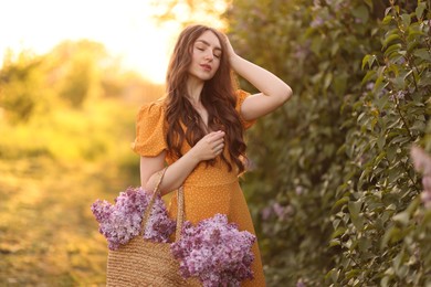 Photo of Beautiful woman with bag of lilac flowers near bush outdoors in morning