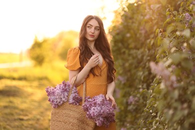 Photo of Beautiful woman with bag of lilac flowers near bush outdoors in morning