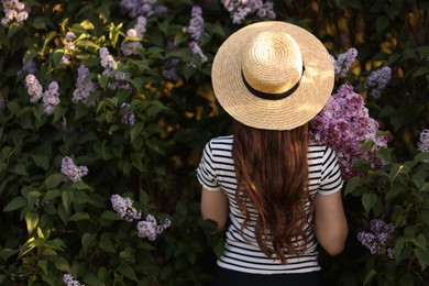 Photo of Woman in straw hat among lilac flowers and leaves outdoors, back view. Space for text