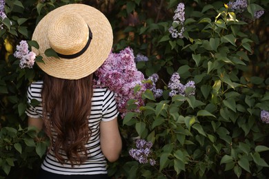 Photo of Woman in straw hat among lilac flowers and leaves outdoors, back view
