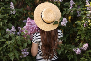 Photo of Woman in straw hat among lilac flowers and leaves outdoors, back view