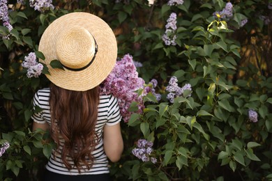 Photo of Woman in straw hat among lilac flowers and leaves outdoors, back view