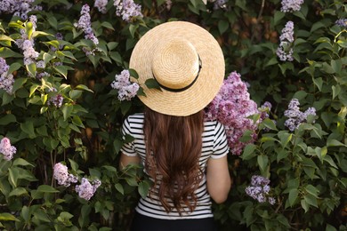 Photo of Woman in straw hat among lilac flowers and leaves outdoors, back view