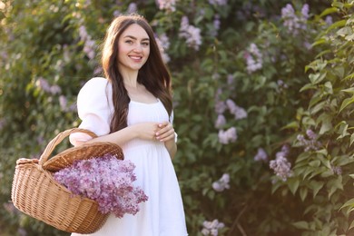 Photo of Smiling woman with basket of lilac flowers near bush outdoors