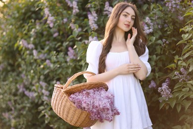 Photo of Beautiful woman with basket of lilac flowers near bush outdoors