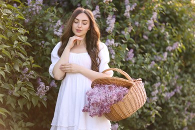 Photo of Beautiful woman with basket of lilac flowers near bush outdoors
