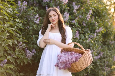 Photo of Beautiful woman with basket of lilac flowers near bush outdoors