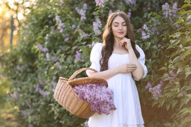 Photo of Beautiful woman with basket of lilac flowers near bush outdoors