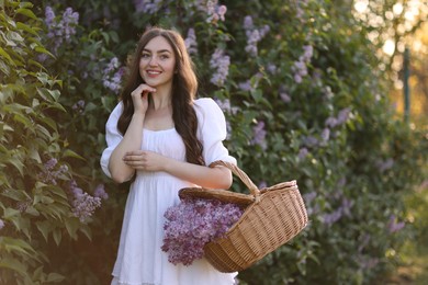 Photo of Smiling woman with basket of lilac flowers near bush outdoors
