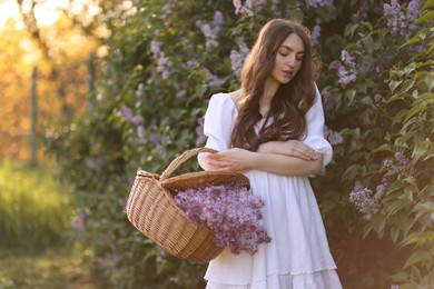 Photo of Beautiful woman with basket of lilac flowers near bush outdoors in morning